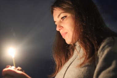 Karanlık, samimi bir ortamda ışıl ışıl parlayan bir maytapa odaklanan bir kadın.