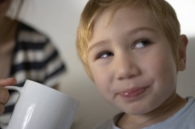 Genç bir çocuk bakıyor, arka planda beyaz bir kupa tutan bir kadın.