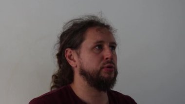 Uzun saçlı sakallı bir adamın portresi beyaz bir arka planda konuşurken yukarı bakıyor.