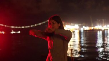 Neşeli genç bir kadın, dışarıda eğleniyor, deniz kenarında dans ediyor, arka planda şehir ışıkları var.