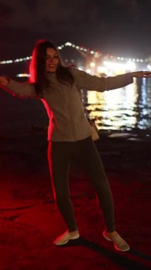 Neşeli genç bir kadın, arka planda şehir ışıklarıyla gece suyun kenarında mutlu bir şekilde dans ediyor.