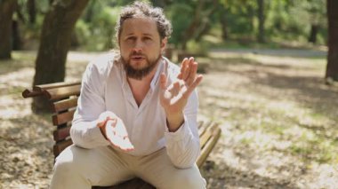 Sakallı ve rastalı ciddi bir adam bankta oturmuş el kol hareketi yapıyor.