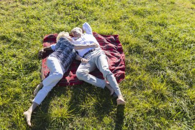 Yaşlı çift, yeşil çimenlerin üzerinde kırmızı bir battaniyeye uzanmış güneşli bir günün tadını çıkarıyorlar.