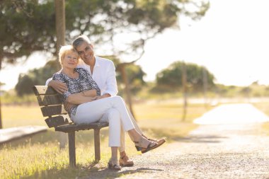 Kıdemli adam ve kadın parkta bir bankta oturuyor, gülümsüyor ve kameraya bakıyor.