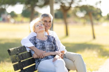 Yaşlı çift parkta bankta dinlenirken, adam kadını arkadan kucaklıyor, mutlu bir anı paylaşıyor.