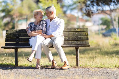 Kıdemli çift bankta oturup kucaklaşıyorlar, dışarıda mutlu ve sevgi dolu bir etkileşimi paylaşıyorlar.