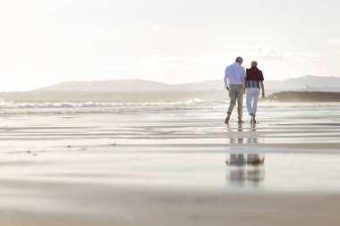 Altın saat boyunca okyanus kıyısında yürüyen mutlu yaşlı çift, birlikte emekliliğin tadını çıkarıyor.