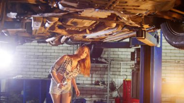 Araba tamir servisi. Araba kaldırma. Genç seksi kadın arabanın altına giriyor ve İngiliz anahtarıyla çalışıyor. Orta çekim