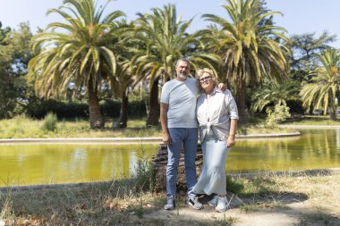 Güneşli bir parkta, palmiye ağaçları ve göleti olan olgun bir çift.