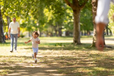 Happy child running towards a parent in a sunny park with an adult in the background
