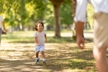 Young boy smiling and running on a sunny day in a green park towards an adult