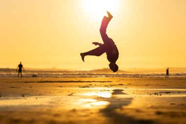 Altın saat boyunca kumlu bir plajda yetenekli bir ters takla atan genç adam.