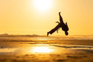 Akrobat plajda sıcak altın bir günbatımına karşı dinamik bir ters takla atıyor.