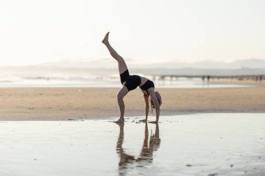 Woman performing a flexible yoga pose with one leg extended upwards on a sandy beach