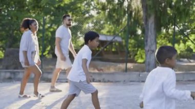 Küçük çocuk sopayla oyun oynamaktan, koşmaktan ve rol yapmaktan zevk alırken, ebeveynler arka planda izliyor.