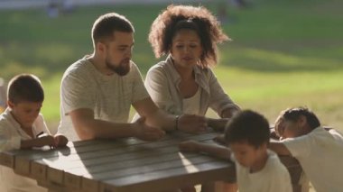 Mutlu bir aile güneşli bir parkta piknik masasında eğleniyor.