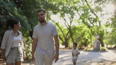 Çocukları onlara koşarken genç ebeveynler parkta yürüyüp konuşuyorlar.