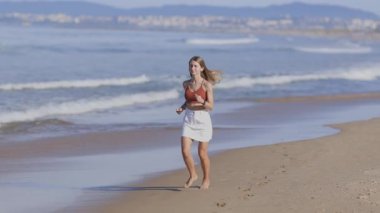 Güzel bir yaz gününün tadını çıkaran mutlu genç bir kadın huzurlu okyanus kıyısında yalınayak koşuyor.