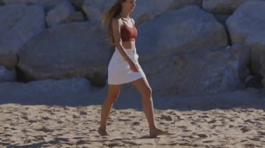Side view of a beautiful young woman with long hair walking barefoot on a sunny sandy beach