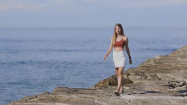 Etek ve bluz giymiş ince bir kadın güneşli bir günde dalgalı bir dalgada yürüyor.