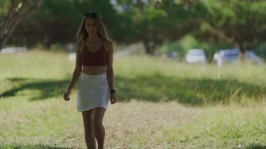Güneşli bir yaz gününde parkta kameraya doğru yürüyen uzun saçlı çekici genç bir kadın.