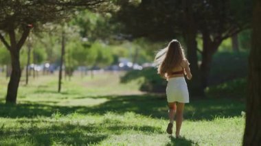 Uzun saçlı genç bir kadın yeşil çimenler ve ağaçlarla çevrili güneşli bir parkta koşuyor.