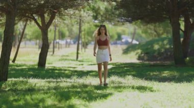 Çekici genç bir kadın, yeşil çimlerle dolu güneşli bir parkta kameraya doğru güvenle yürüyor.
