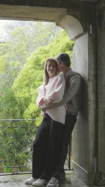 Genç çift balkonda samimi bir anı paylaşıyor, tarihi bir binayla kucaklaşıyorlar ve arka planda yemyeşil bir alan var.