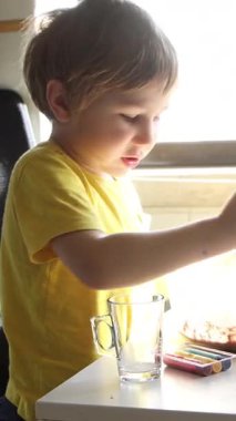 Sarı tişörtlü çocuk, cam kasedeki malzemeleri beyaz masada karıştırıyor.