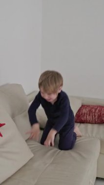 Genç çocuk eğleniyor, zıplıyor ve parlak bir oturma odasında rahat bir kanepede oynuyor.