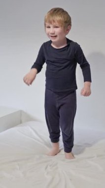 Pijama giyen sarışın çocuk beyaz yatakta neşeyle dans ediyor.