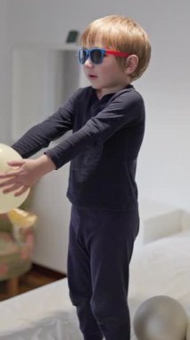 Yatakta balonlarla oynayan güneş gözlüklü sarışın çocuk.