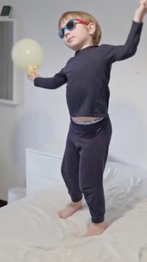 Güneş gözlüğü takan bir çocuk yatakta atlarken beyaz bir balonla oynar.