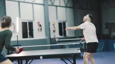 Genç adam ve kadın yoğun bir şekilde bir spor salonunda masa tenisi oynuyorlar, adam puanı kazanıyor ve kutluyor.