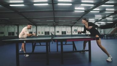 Kapalı bir spor salonunda masa tenisi oynayan odaklanmış genç adam ve kadın.