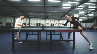 Genç adam ve kadın yoğun bir şekilde büyük bir salonda masa tenisi oynuyorlar.