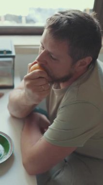 Üzgün adam mutfak zemininde oturmuş elma yiyor.