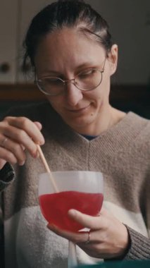 Kadın kırmızı sıvıyı bir kasede karıştırıyor. Mutfakta tahta bir sopayla oturuyor.