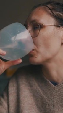 Gözlük takan ve günlük kıyafetler giyen bir kadın mutfaktaki plastik bardaktan su içiyor.