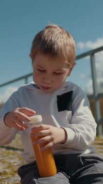 Çocuk dışarıda, korkuluk ve nehrin yanında bir şişe meyve suyu açmaya çalışıyor.