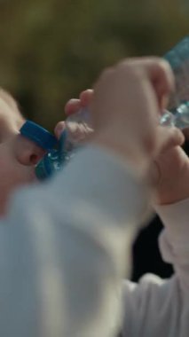 Parktaki plastik bir şişeden su içen genç bir çocuk, annesi onu izliyor.