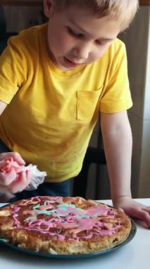 Genç çocuk bir ev mutfağında pembe kremayla pasta süslüyor.