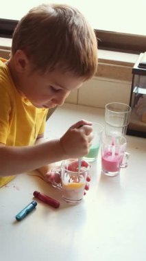 Genç bir çocuk renkli sıvılarla deney yapıyor, onları beyaz bir masada bir bardakta karıştırıyor.