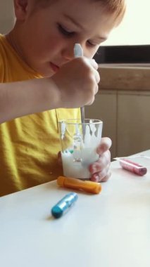 Genç bir çocuk beyaz bir masada renkli bir macunu bir bardak süte karıştırıyor.