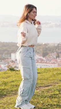 Enerjik dansçı çimenli tepe üzerinde manzaralı nehir ve şehir planıyla çeşitli hareketler yapar.