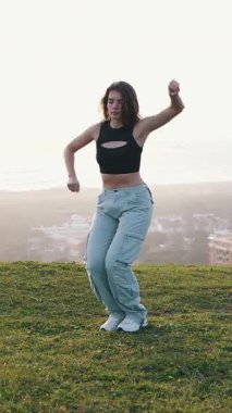 Dansçı kadın, arka planda şehir manzarası ve günbatımı olan otlu bir tepede çeşitli dans hareketleri sergiliyor.