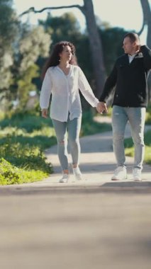 Genç bir çift güneşli bir parkta rahatça geziniyor, el ele tutuşuyor ve bir anlık yakınlık kuruyorlar.