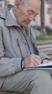 Kıdemli adam parkta boş zamanlarının tadını çıkarıyor, kulaklıkla müzik dinlerken bir deftere yazı yazıyor.