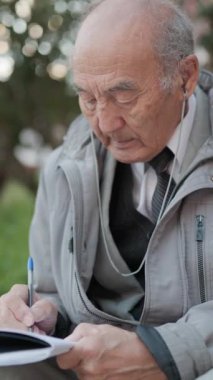 Kıdemli adam bir parkta bankta otururken defterine not alıyor.