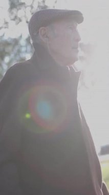 Kahverengi paltolu ve düz şapkalı yaşlı adam güneşli bir parkta geziniyor.